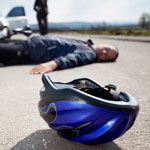a photo depicting a byciclying accident, where in the foreground a helment lays on the pavement, while in the background the bicylist lays on the groud in front of a car.